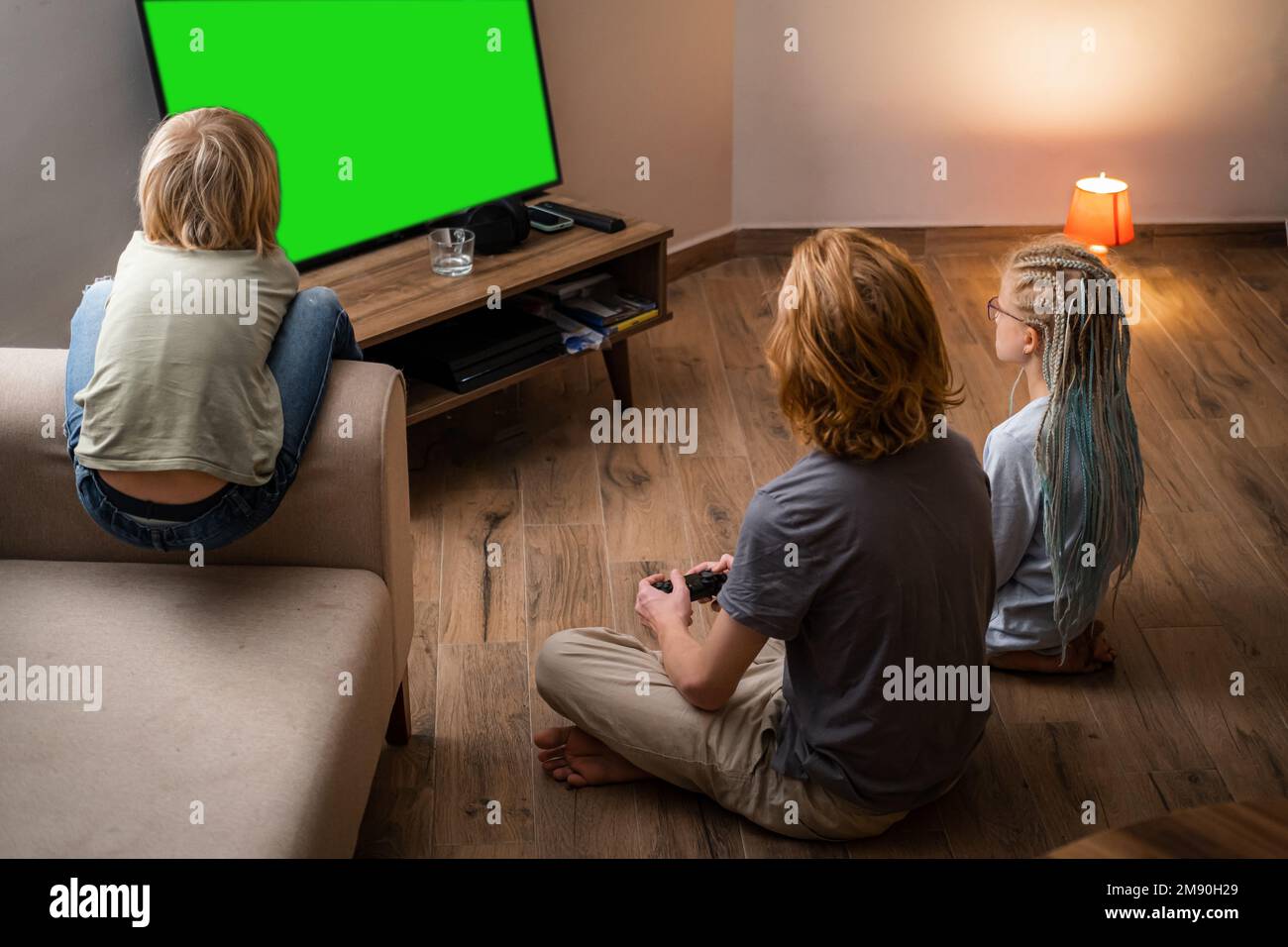 Trois enfants jouant au jeu vidéo Green Screen chromakey sur la console de télévision à l'aide de la manette tout en étant assis à la maison, frères et soeur assis sur le sol Banque D'Images