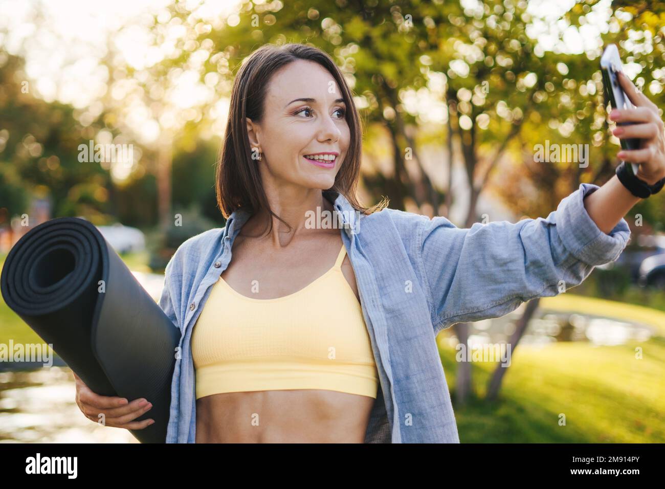 Femme adulte souriante dans des vêtements de sport debout dans un parc herbeux prenant un selfie au téléphone après avoir fait du yoga. FIT femme jogging à l'extérieur. Un style de vie heureux Banque D'Images