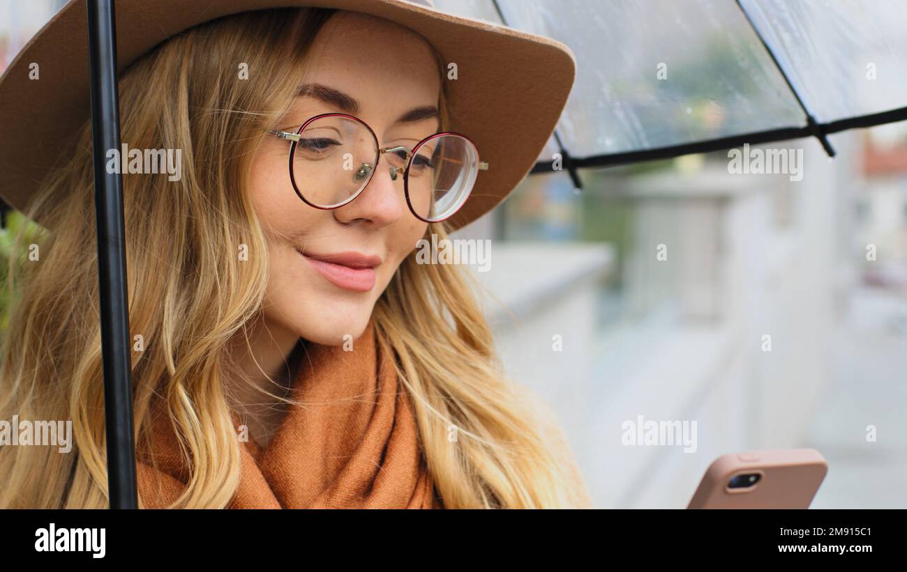 Gros plan Femme caucasienne souriante sous parapluie avec téléphone extérieur dame tenant smartphone fille avec chapeau et lunettes discutant avec mobile Banque D'Images