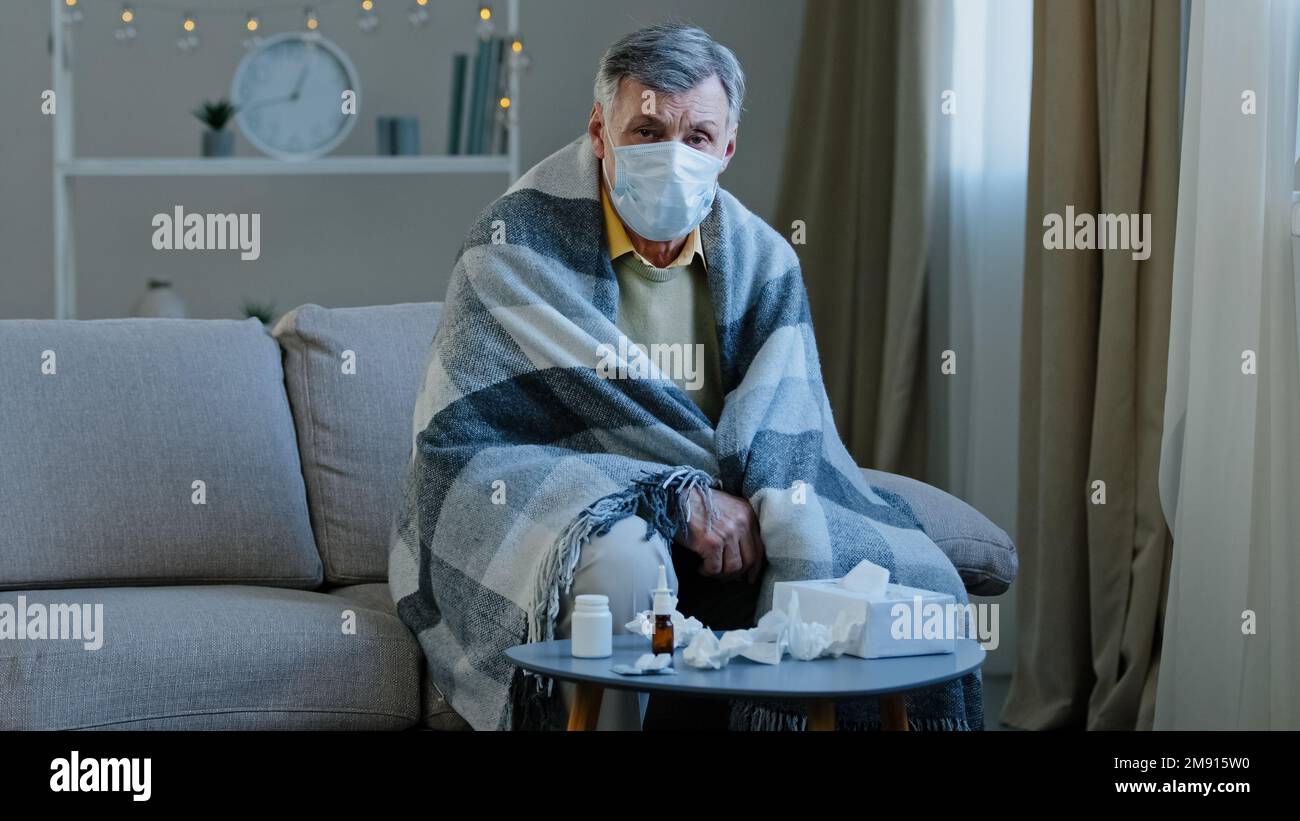 Un grand-père malade contrarié dans un masque médical assis sur un canapé un homme âgé caucasien enveloppé dans une couverture se sent froid un aîné malade mature regardant les symptômes de la caméra Banque D'Images