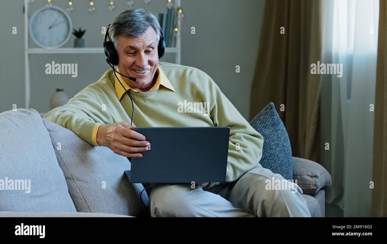 Homme de conversation âgé de 60s ans de race blanche travaillant à distance de la maison assis sur un canapé dans un casque parlant avec un ordinateur portable de webcam. Entraîneur en ligne de 60s anciens enseignants Banque D'Images