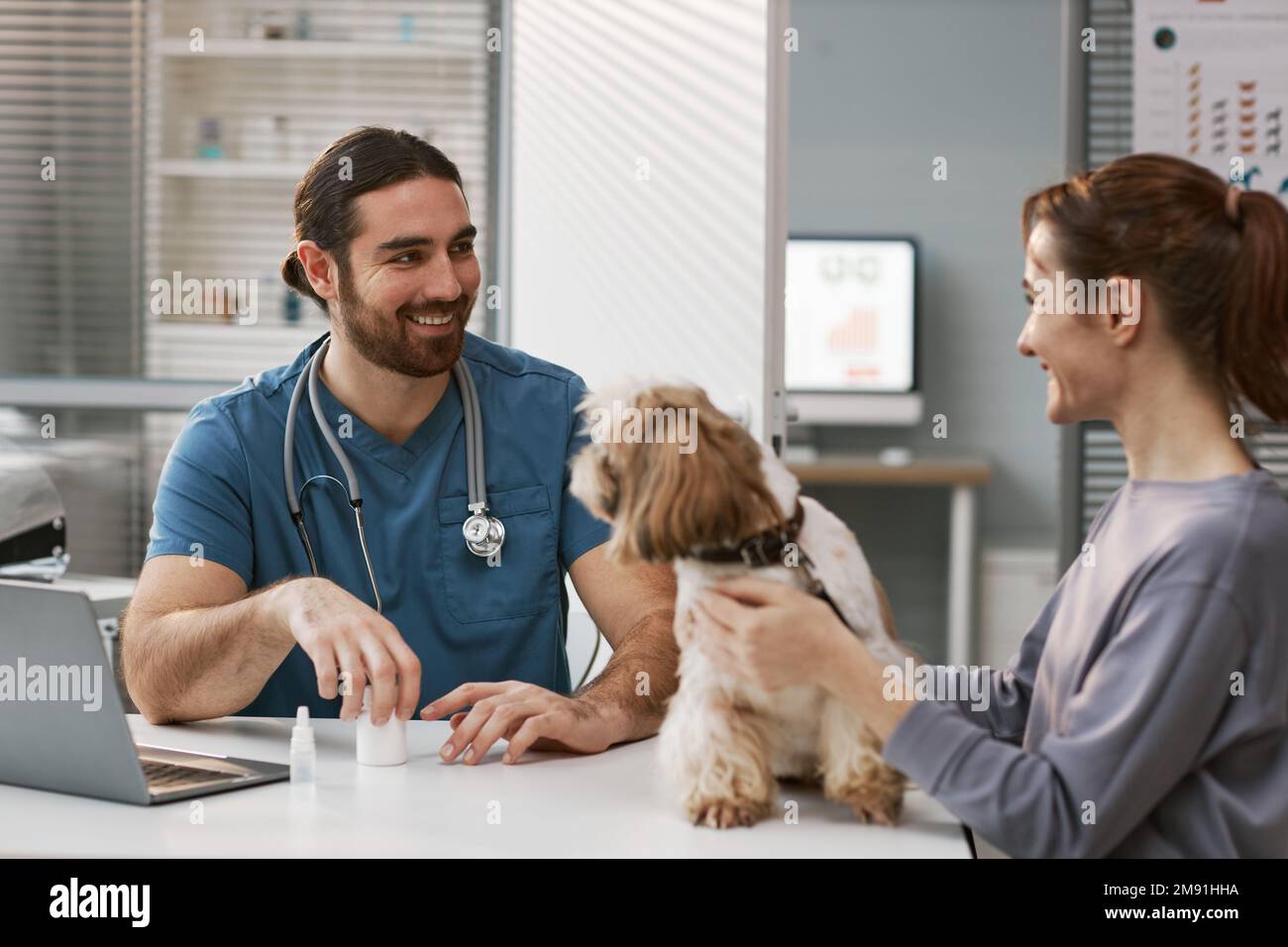 Joyeux jeune vétérinaire masculin en uniforme regardant la cliente féminine avec son animal de compagnie tout en la consultant et en prescrivant des médicaments pour l'animal malade Banque D'Images