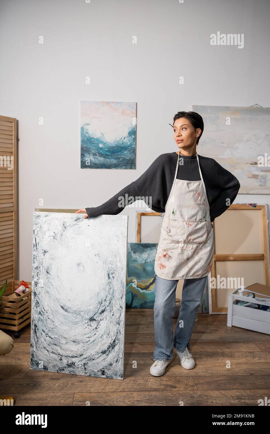 Artiste aux cheveux courts debout avec la main sur la hanche près de la peinture dans l'atelier Banque D'Images