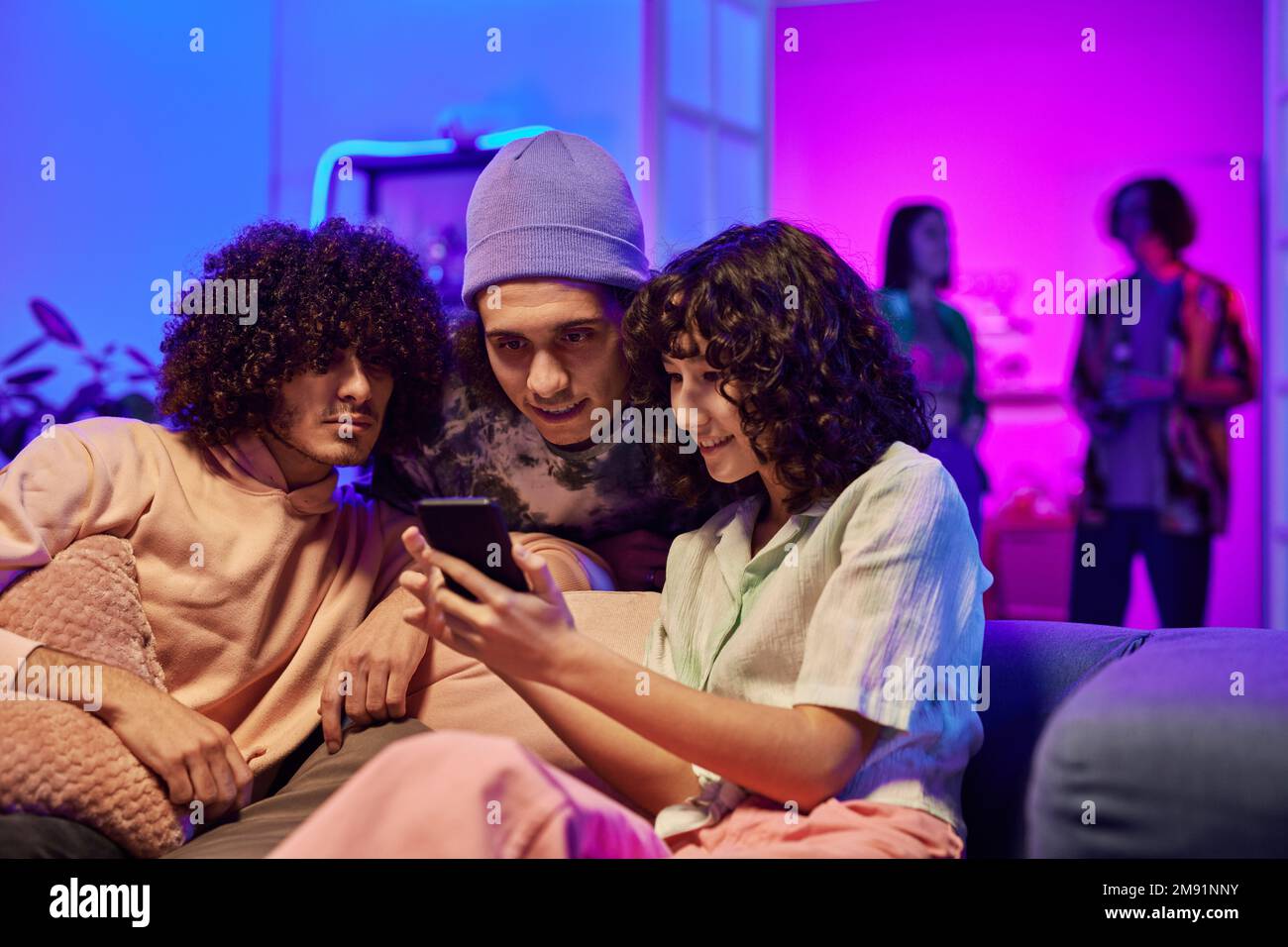 Jeune jolie femme aux cheveux frisés foncés et ses deux amis masculins interculturels regardant la vidéo en ligne sur un smartphone lors d'une fête à la maison Banque D'Images