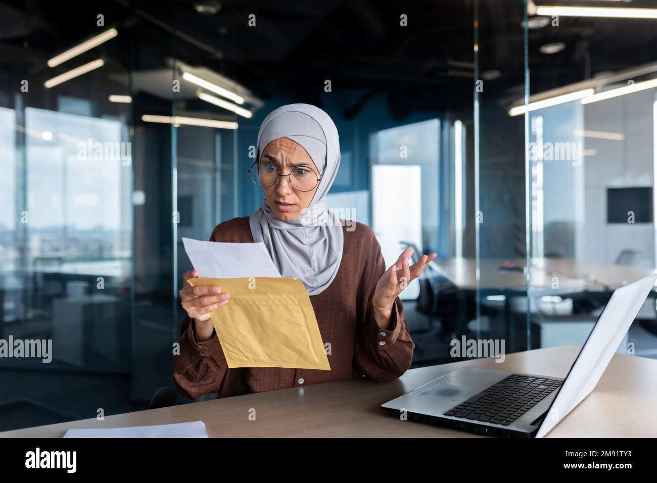 Une femme d'affaires contrariée dans le hijab travaillant à l'intérieur du bureau, une femme a reçu un courrier d'enveloppe avec de mauvaises nouvelles, un employé de bureau contrarié par une notification travaillant sur le lieu de travail avec un ordinateur portable. Banque D'Images