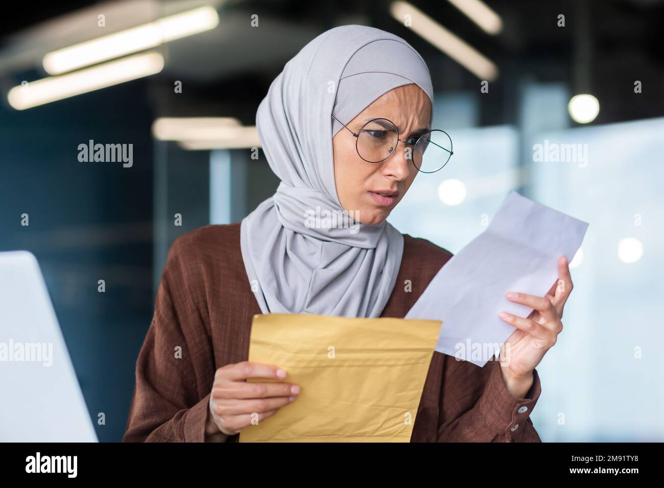 Une femme d'affaires contrariée dans le hijab travaillant à l'intérieur du bureau, une femme a reçu un courrier d'enveloppe avec de mauvaises nouvelles, un employé de bureau contrarié par une notification travaillant sur le lieu de travail avec un ordinateur portable. Banque D'Images