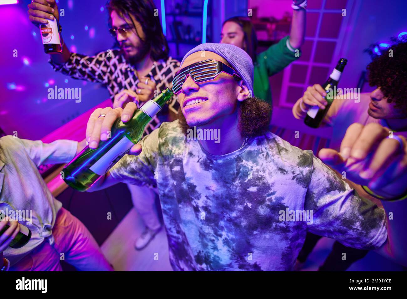 Joyeux jeune homme avec une bouteille de bière dansant entre amis interculturels ecstatiques appréciant la fête à la maison le week-end ou vendredi soir Banque D'Images