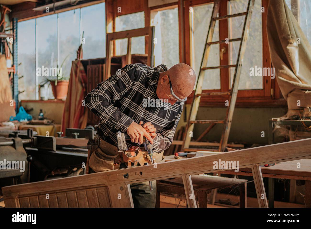 Ouvrier du bois âgé dans des lunettes de protection, en train de percer des planches de bois tout en travaillant dans la menuiserie Banque D'Images