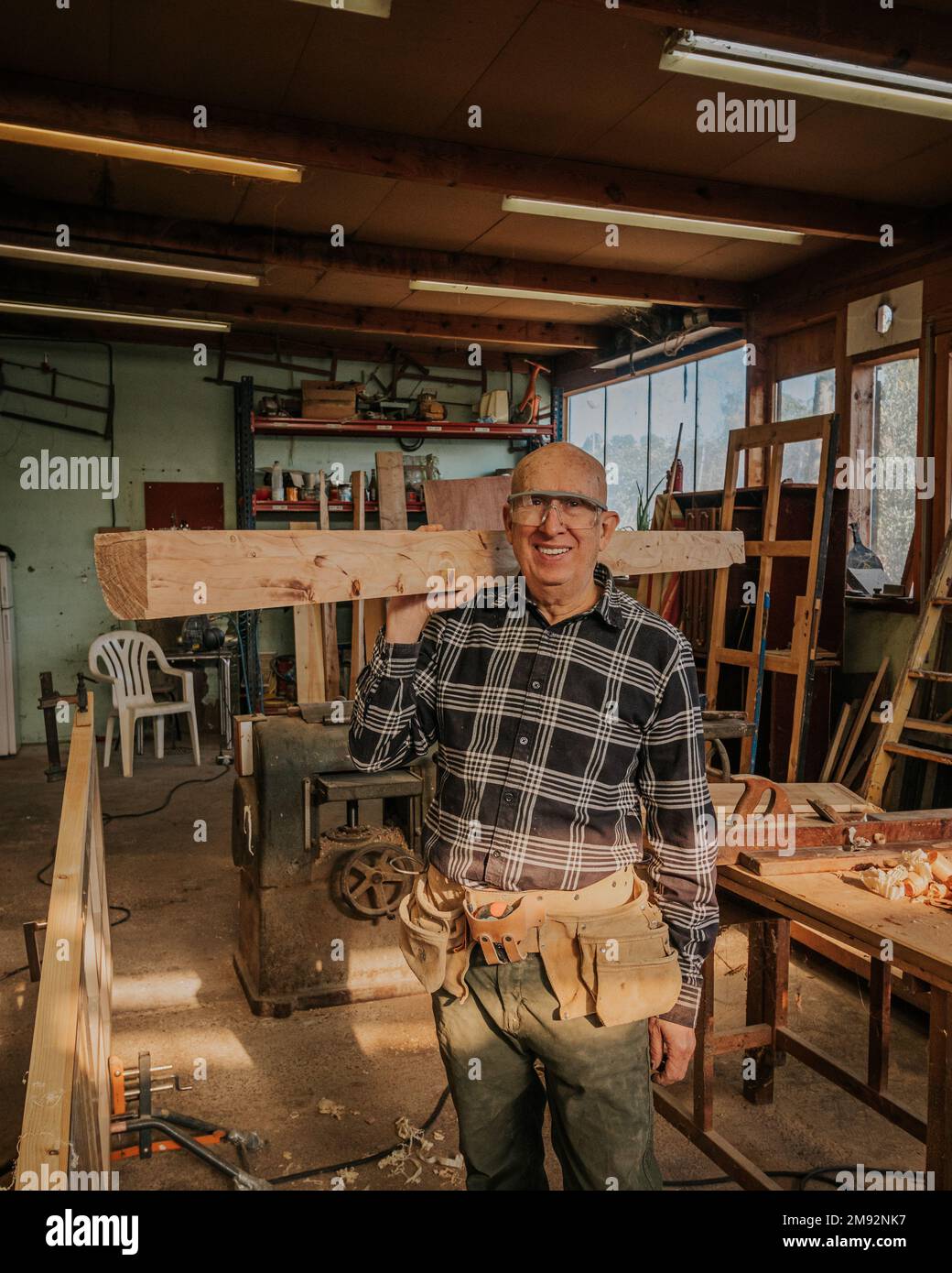 Menuisier mâle senior souriant dans des lunettes de protection transportant du bois sur l'épaule dans la menuiserie professionnelle pendant le processus de réparation Banque D'Images