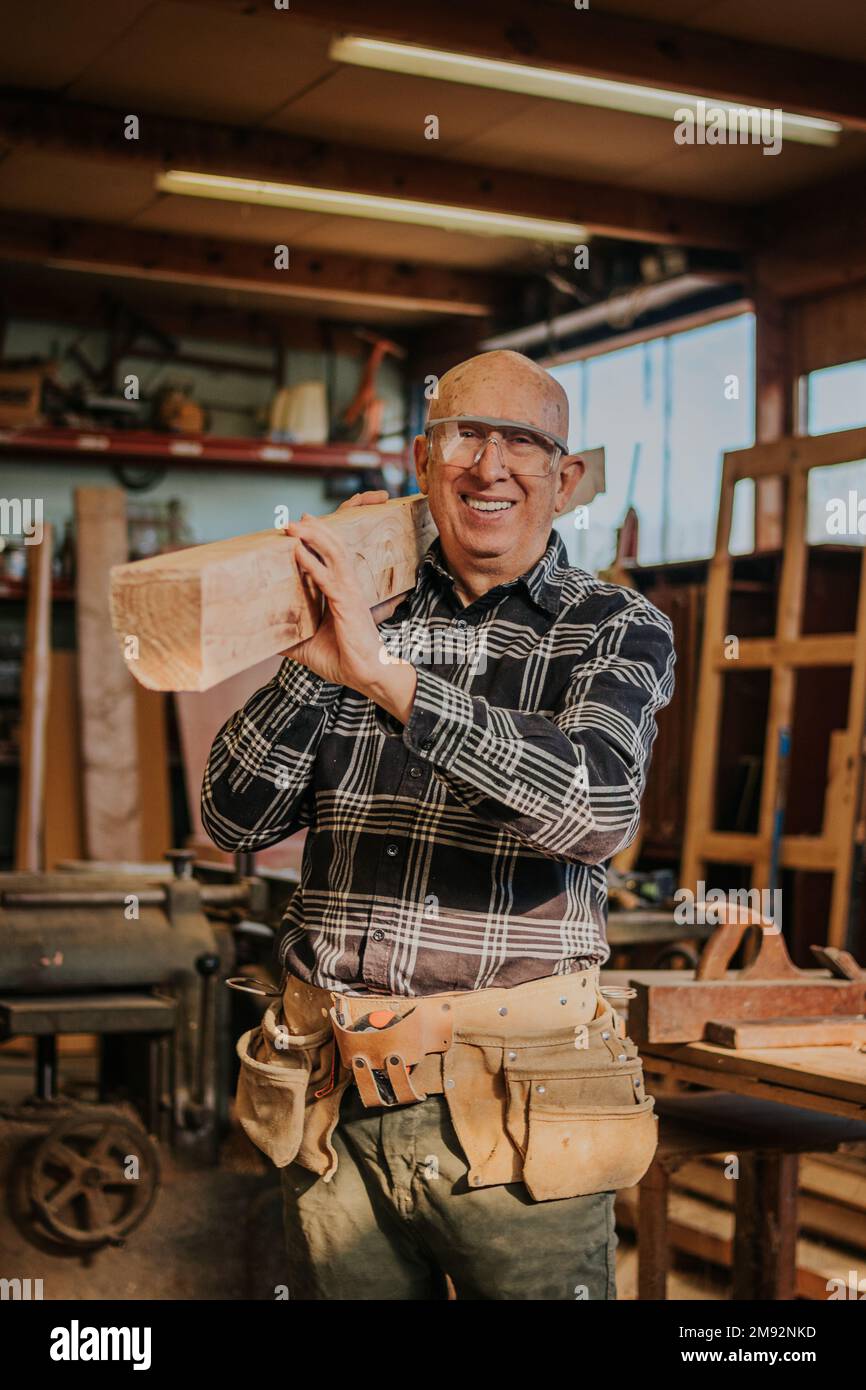 Menuisier mâle senior souriant dans des lunettes de protection transportant du bois sur l'épaule dans la menuiserie professionnelle pendant le processus de réparation Banque D'Images