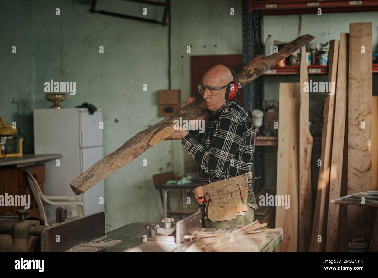 Menuisier mâle dans des lunettes de protection transportant du bois sur l'épaule dans la menuiserie professionnelle pendant le processus de réparation Banque D'Images