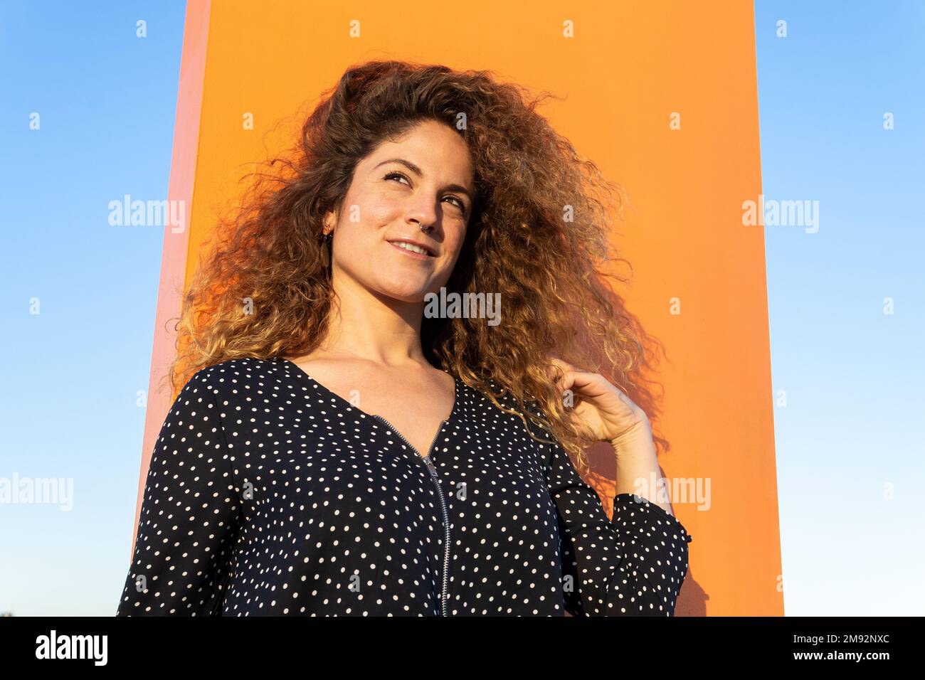 De dessous, jeune femme souriante dans une chemise à pois touchant les cheveux bruns bouclés tout en se tenant contre le ciel bleu dans le vent Banque D'Images