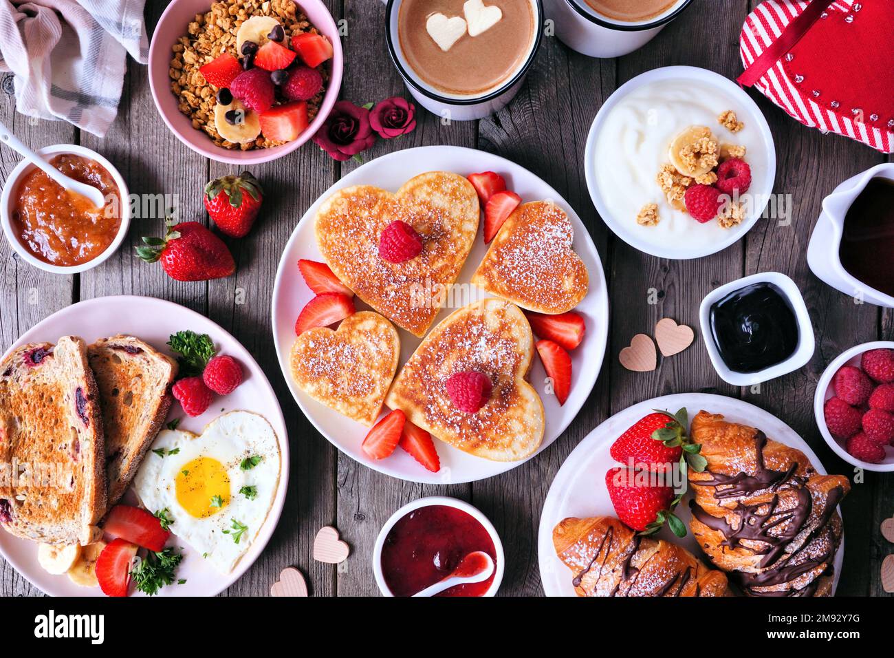 Scène de table pour le brunch de la Saint-Valentin ou de la fête des mères. Vue de dessus sur un fond en bois sombre. Crêpes en forme de cœur, œufs et assortiment de plats sur le thème de l'amour. Banque D'Images