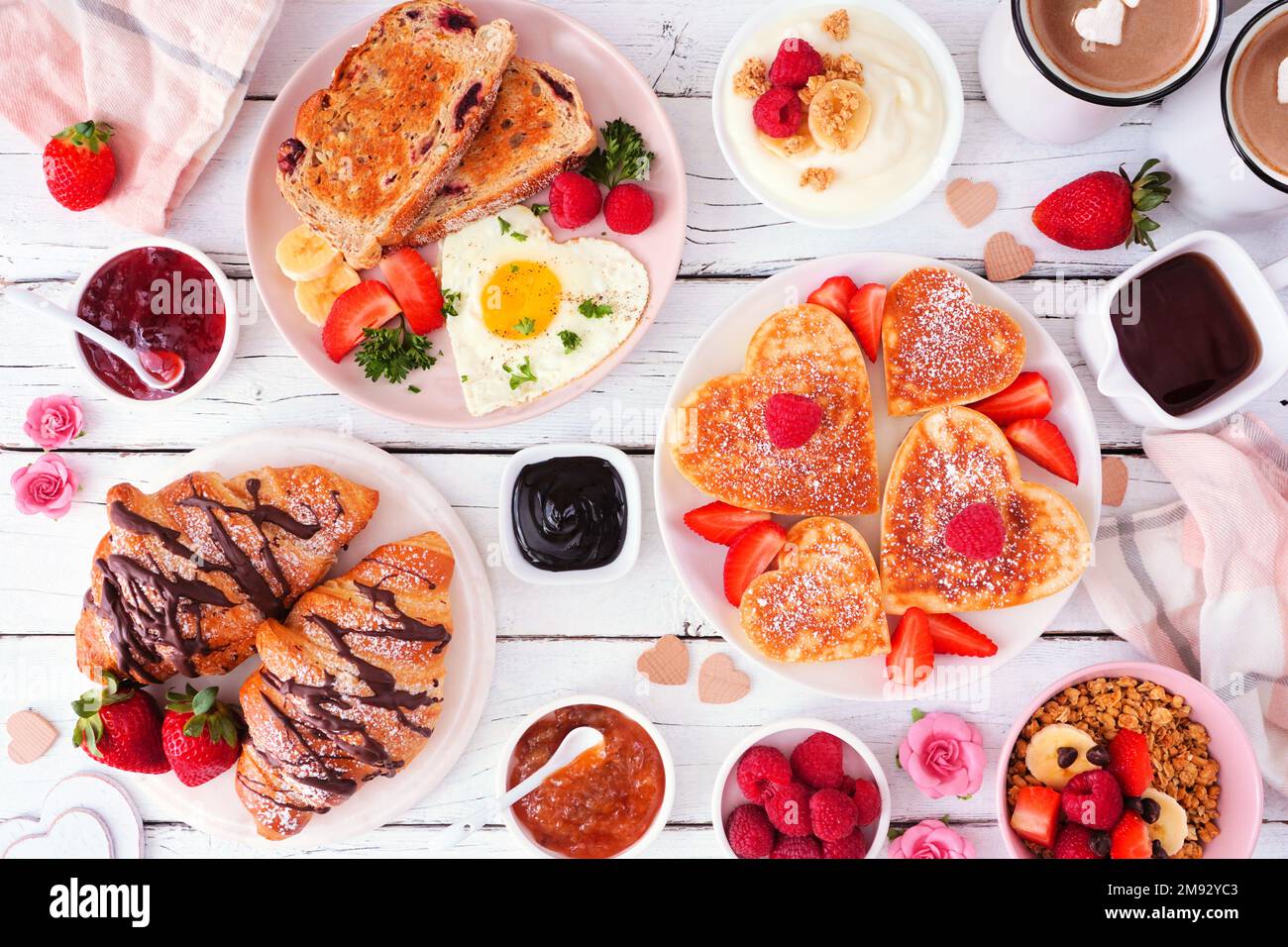 Scène de table pour le brunch de la Saint-Valentin ou de la fête des mères. Vue en hauteur sur fond de bois blanc. Crêpes en forme de cœur, œufs et plats assortis sur le thème de l'amour. Banque D'Images