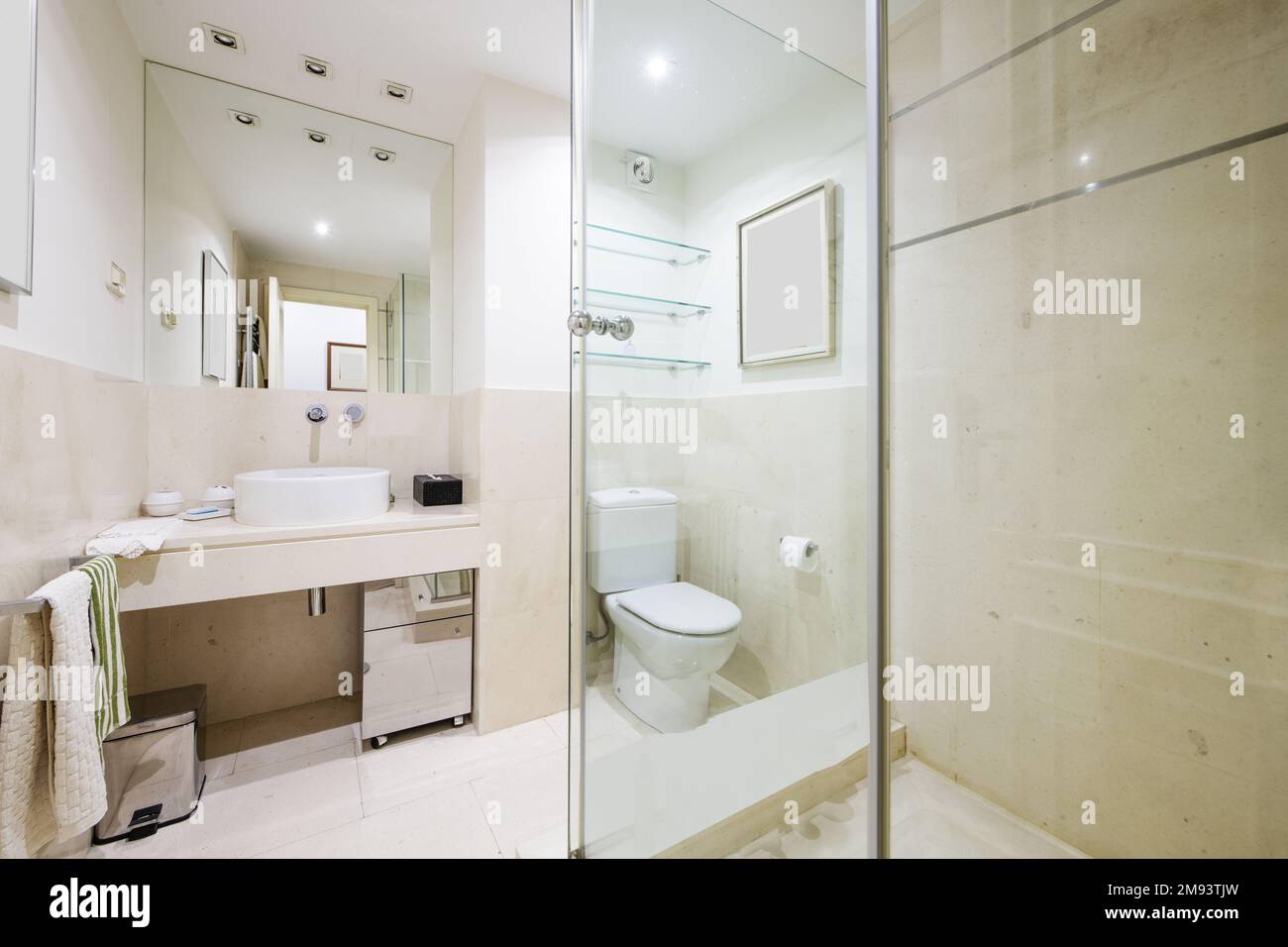 Belle salle de bains avec carrelage en marbre blanc avec miroir intégré au mur et toilettes en porcelaine, appliques murales dans le faux plafond, étagère en verre Banque D'Images