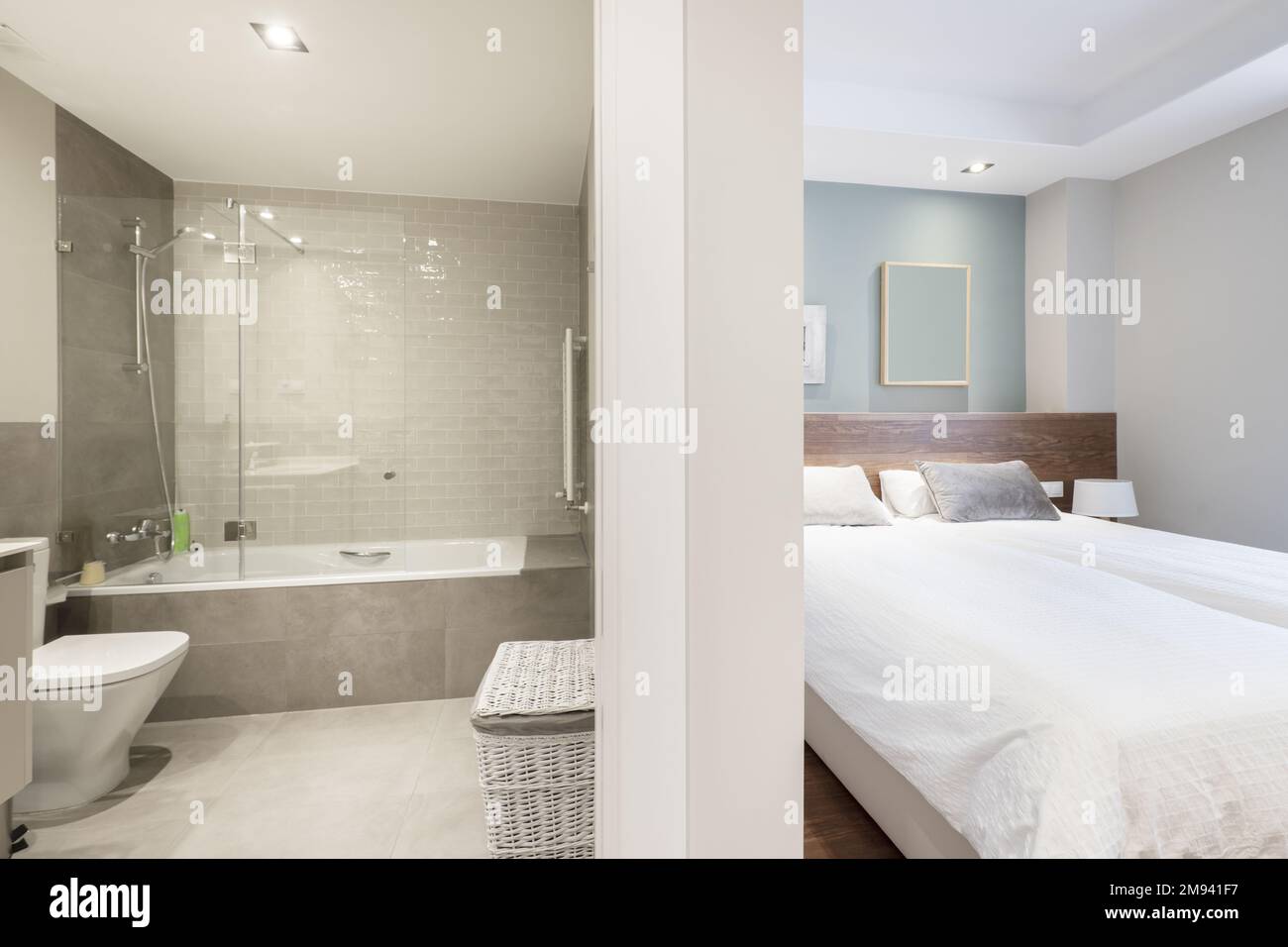 Salle de bain avec baignoire avec cloisons en verre, carrelage gris et chambre avec lit double avec couette blanche Banque D'Images