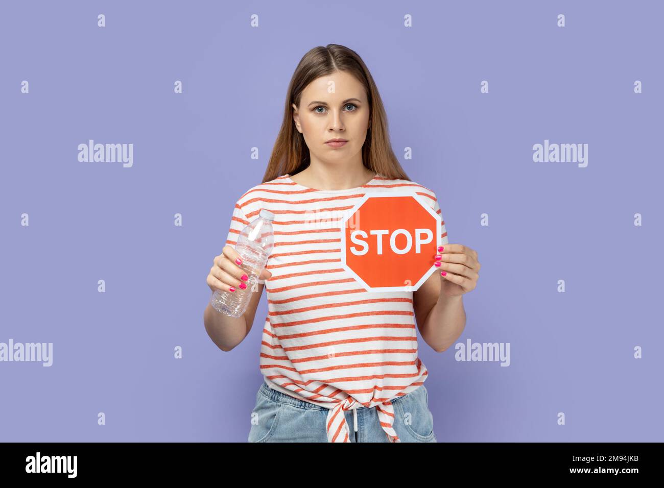 Portrait d'une jeune femme blonde sérieuse et très stricte portant un T-shirt rayé avec un panneau stop rouge et une bouteille en plastique dans les mains. Studio d'intérieur isolé sur fond violet. Banque D'Images