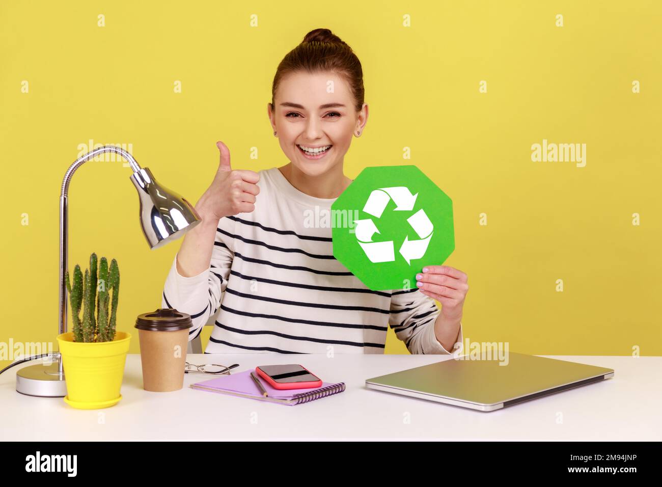 Responsable positif souriant bonne femme tenant main l'affiche de recyclage verte et montrant le pouce vers le haut, assis sur le lieu de travail avec un ordinateur portable. Studio d'intérieur tourné isolé sur fond jaune. Banque D'Images