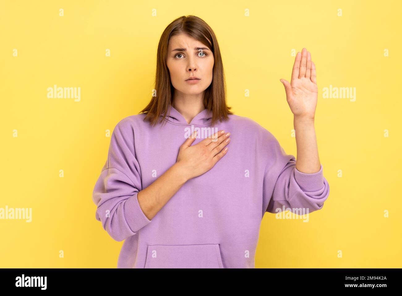 Femme consciente sérieuse tenant une main sur la poitrine et élevant une autre attachante, confiance et honnête, patriotisme, portant le pull à capuche violet. Studio d'intérieur isolé sur fond jaune. Banque D'Images