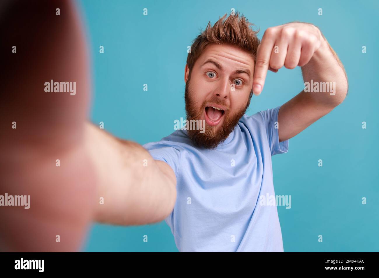 Portrait de l'homme barbu prenant le selfie ou faisant un appel vidéo, regardant la caméra POV, point de vue de la photo, pointant le doigt vers le bas, souscrire. Studio d'intérieur isolé sur fond bleu. Banque D'Images