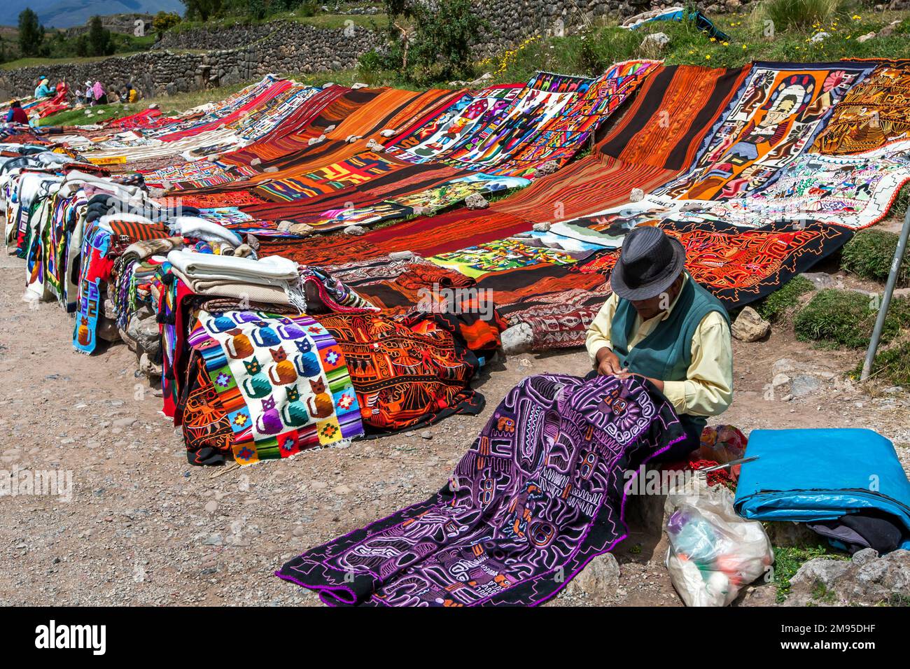 Un homme piquant un tapis péruvien à la périphérie de Cusco au Pérou. En arrière-plan se trouve une variété colorée de tapis à la vente. Banque D'Images