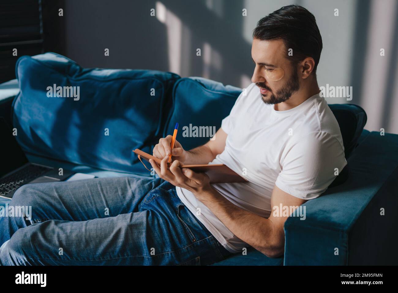Jeune homme de rêve portant un t-shirt pour écrire des souvenirs dans un carnet de notes, assis sur un canapé bleu, restez à la maison et passez du temps libre dans le salon Banque D'Images