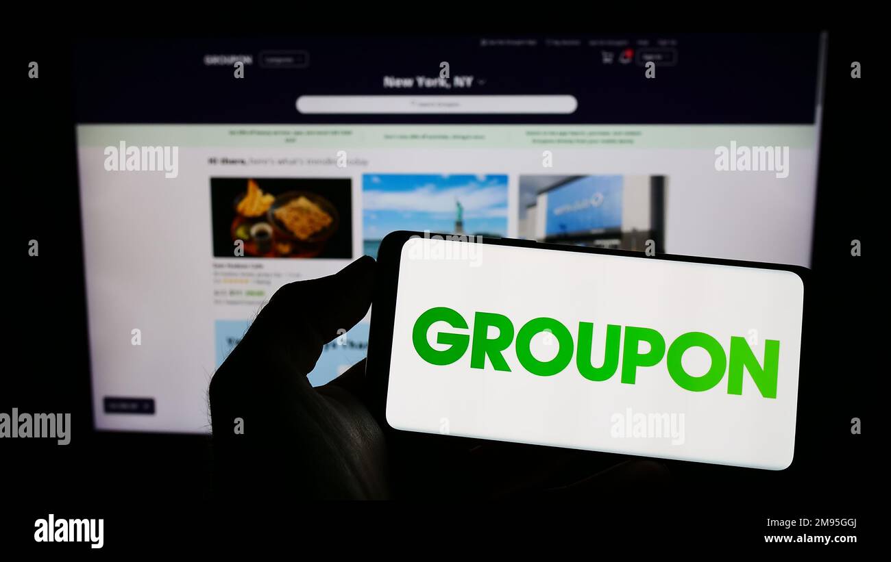 Personne tenant un téléphone mobile avec le logo de la société américaine de commerce électronique Groupon Inc. À l'écran en face de la page Web d'affaires. Mise au point sur l'affichage du téléphone. Banque D'Images