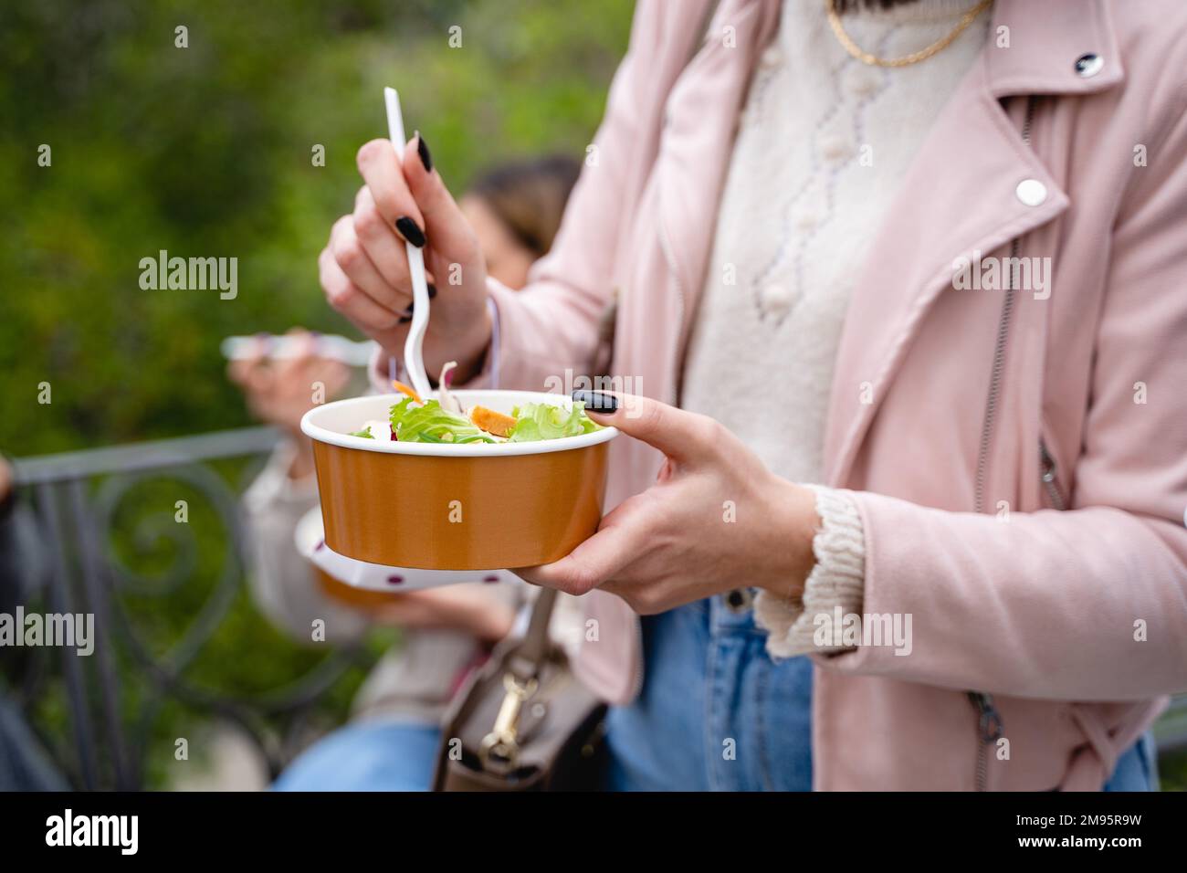 Gros plan sur les mains d'une jeune femme qui apprécie une salade saine dans un bol en papier recyclé. Vêtue d'une veste en cuir rose tendance, elle se tient debout dans une pub Banque D'Images