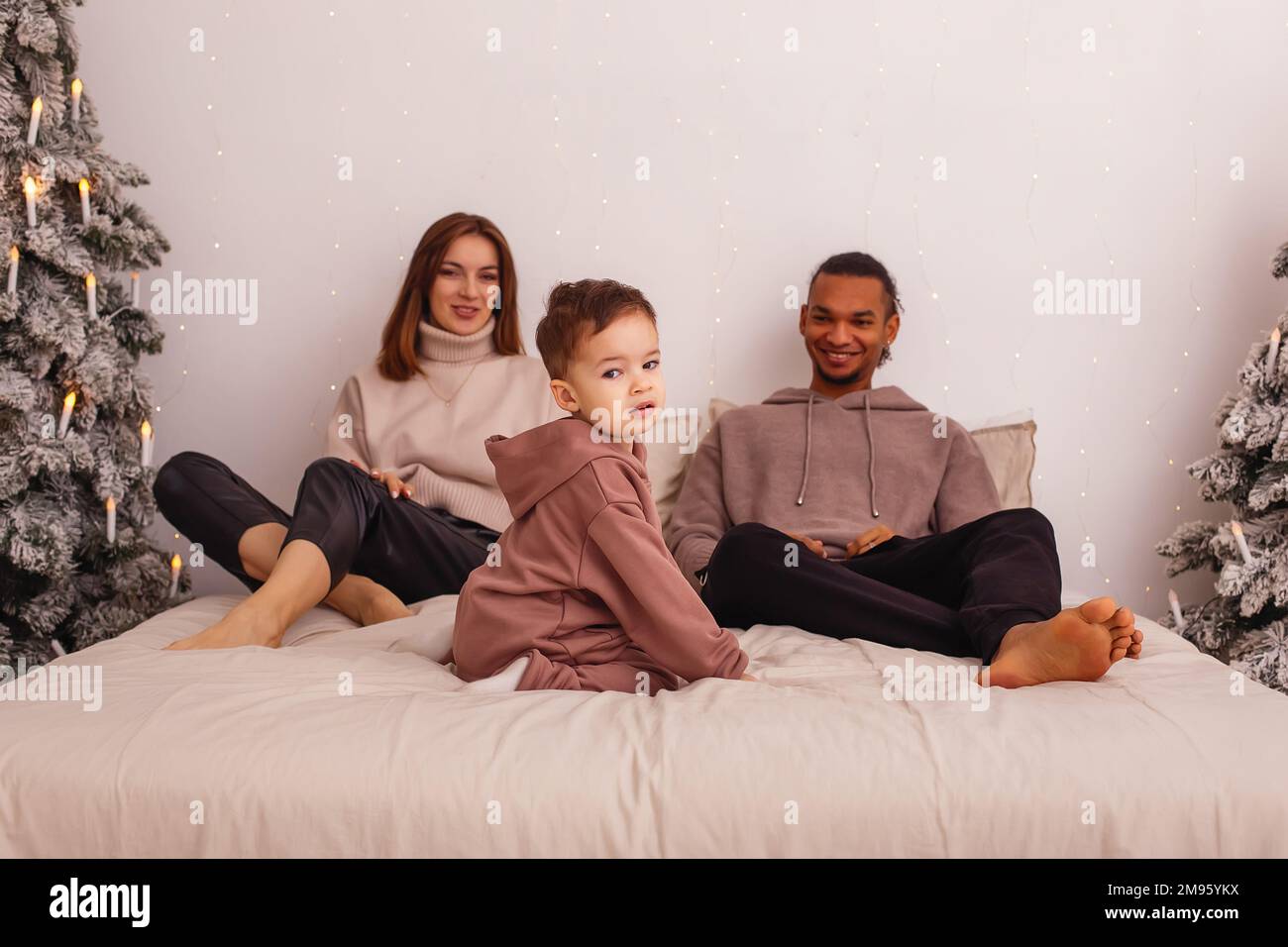 Bonne famille multiraciale avec un petit enfant, assis sur le lit, près de l'arbre de Noël Banque D'Images