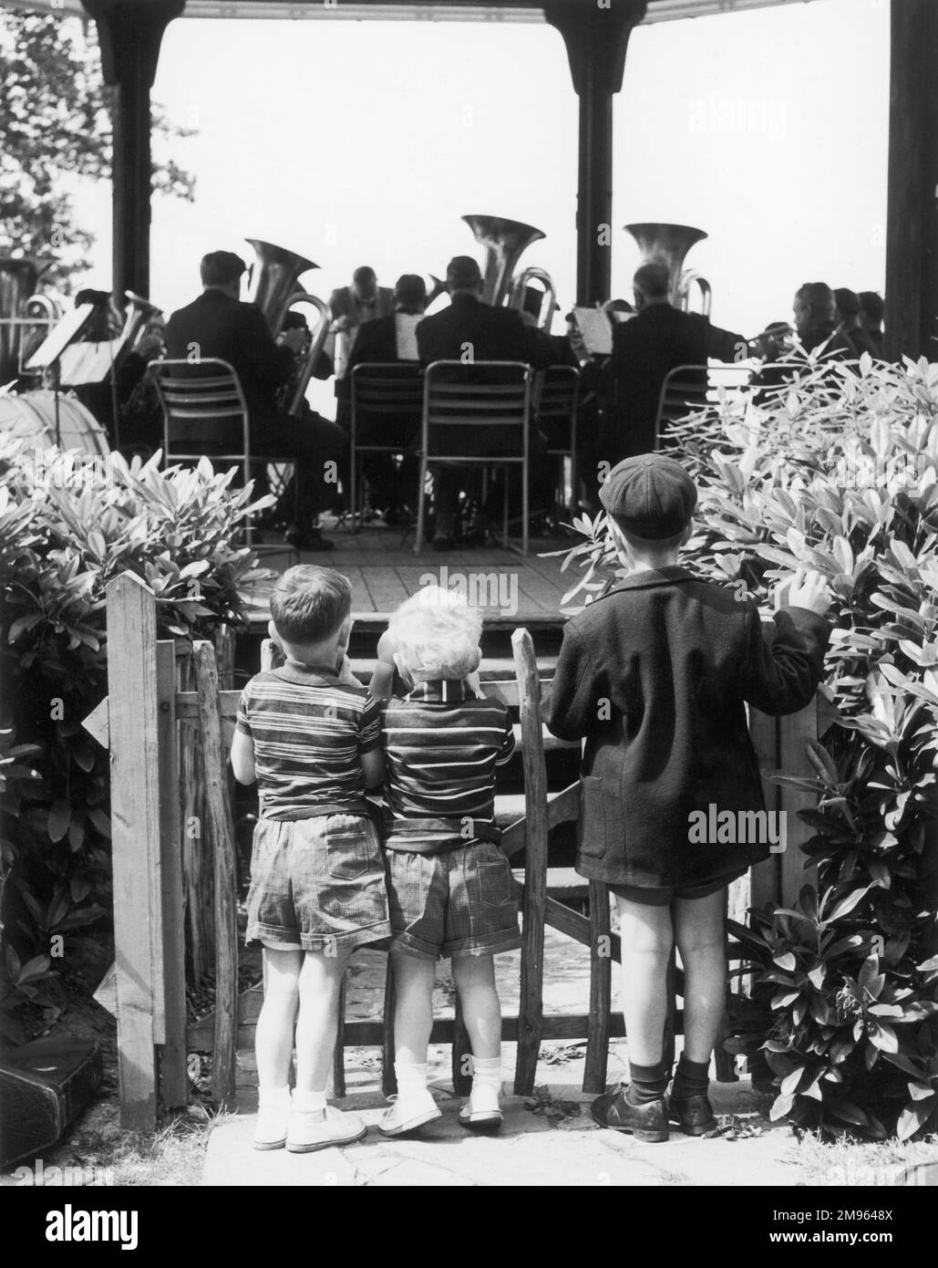 Trois petits garçons se tiennent derrière une porte de piquetage et regardent un groupe de cuivres jouer dans un kiosque. Banque D'Images