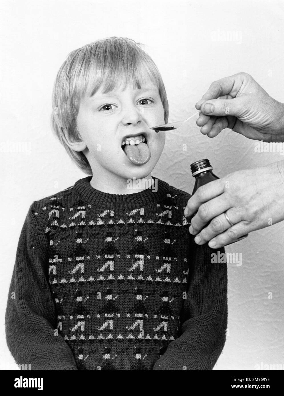 Un petit garçon prenant à contrecœur son médicament -- quelqu'un vient du verser d'une bouteille dans une cuillère pour lui. Il colle sa langue dans le dégoût. Banque D'Images