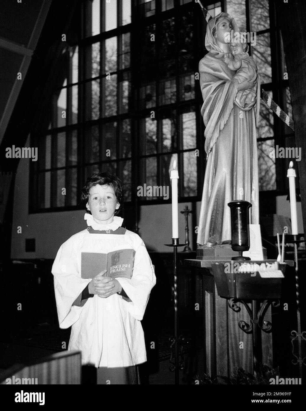 Un choirboy chantant solo devant un vitrail moderne à l'église Salfords, Horley, Surrey. Il tient un livre intitulé "louange anglais". Il y a une statue de la Vierge Marie et de l'enfant à côté de lui. Banque D'Images