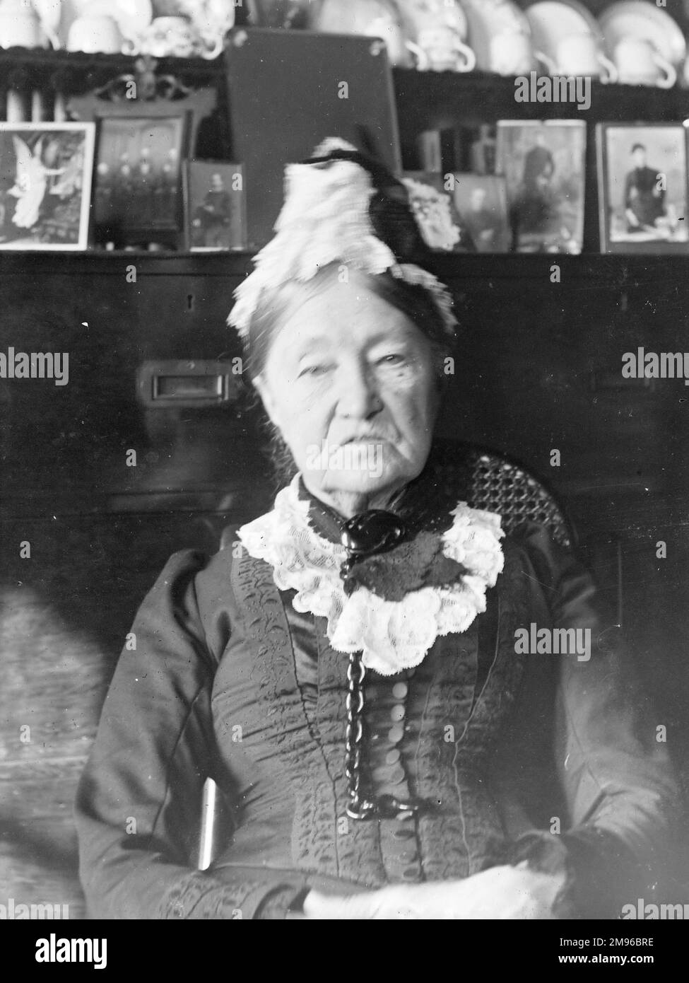 Une femme âgée en costume de style victorien assise devant une commode galloise, au centre du pays de Galles. La commode contient des photos de famille et de la vaisselle. Banque D'Images