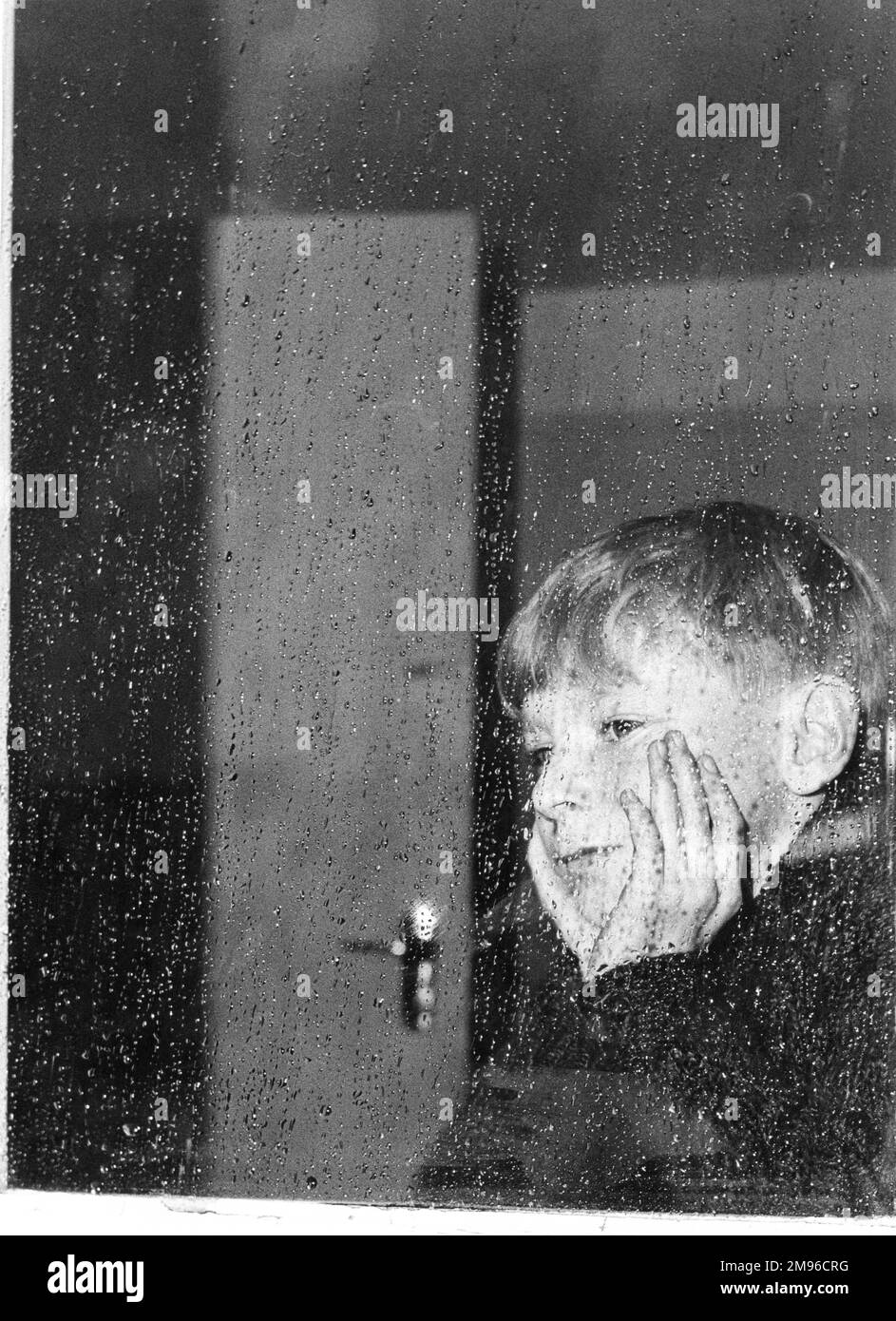 Un petit garçon ennuyé pairs à travers un panneau de fenêtre le jour de la pluie. Banque D'Images