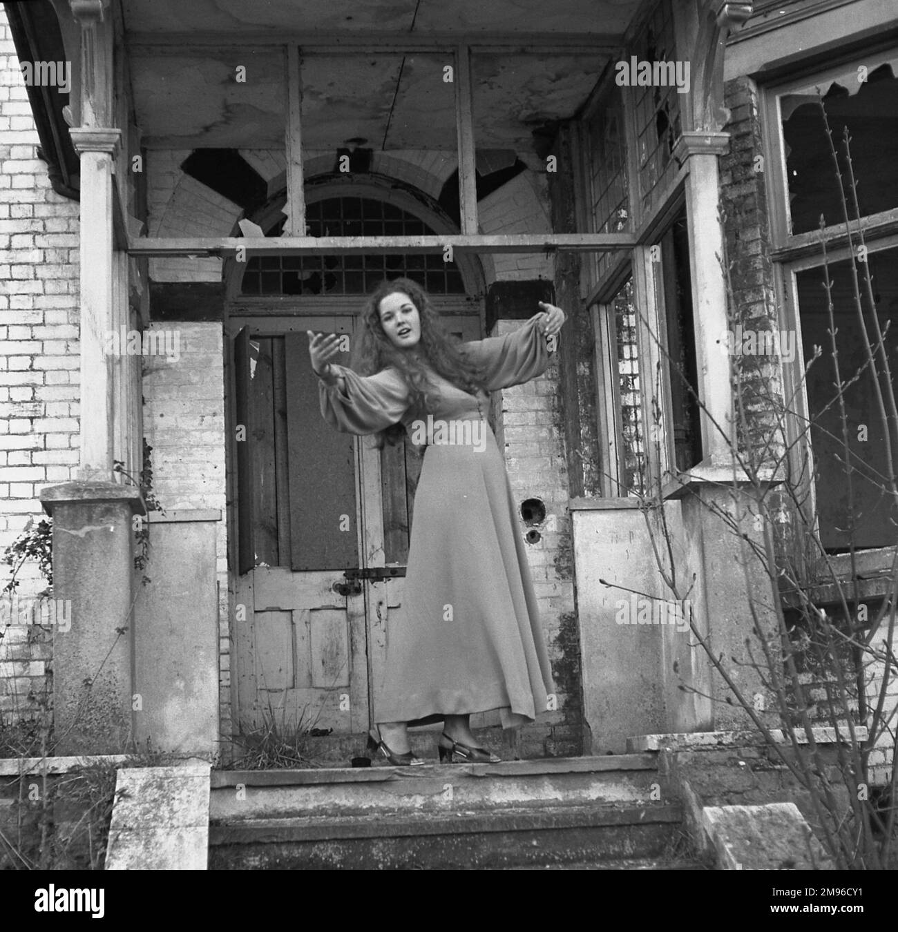 Une jeune femme dans une longue robe pose artistiquement à la porte d'une maison abandonnée avec des fenêtres cassées. Banque D'Images