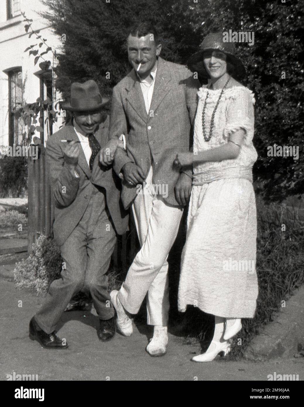 Deux hommes et une femme, posant pour leur photo bras-dans-bras à l'extérieur d'une maison. L'homme de gauche frappe une posture drôle avec ses genoux qui se cognent ensemble. Banque D'Images