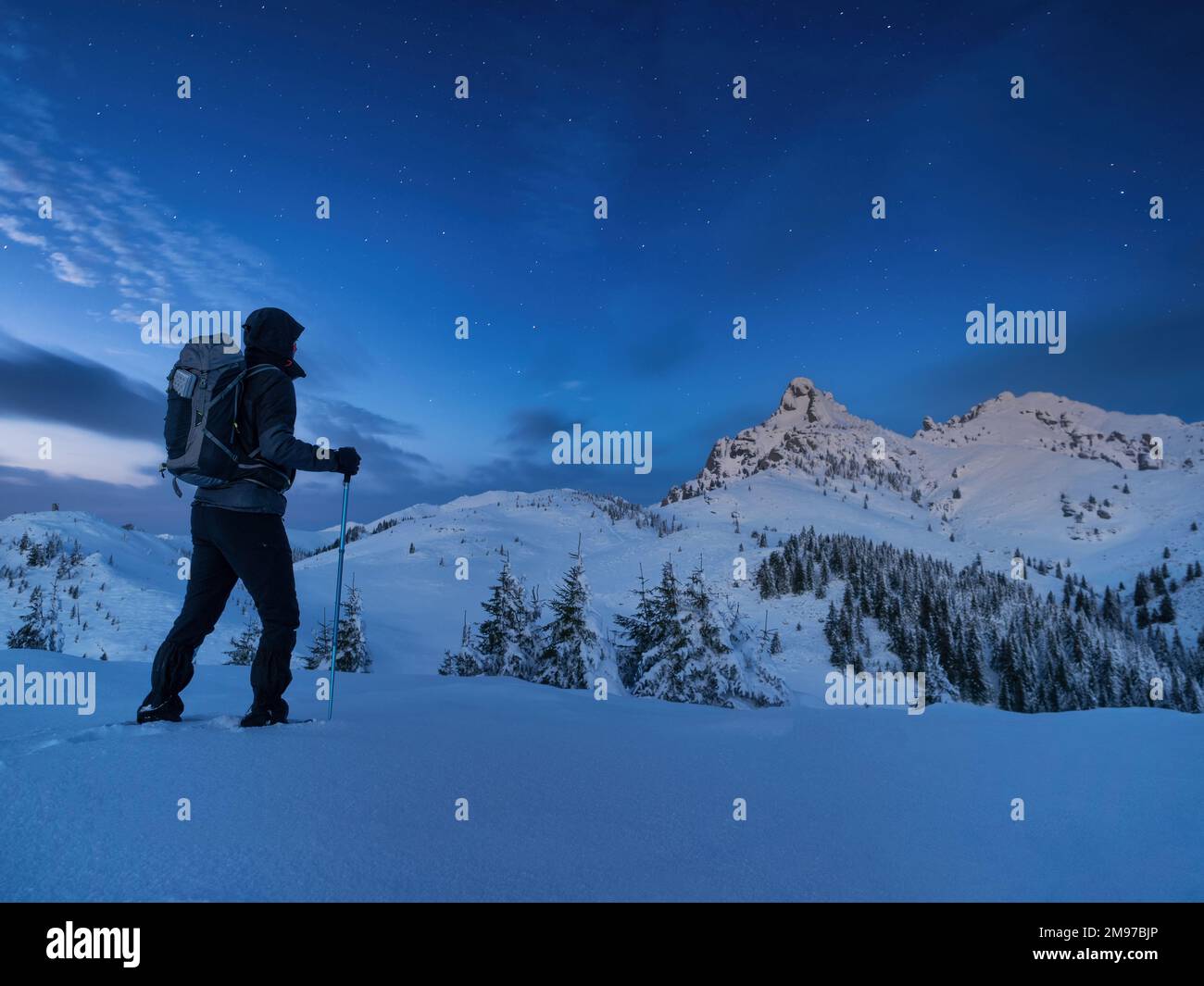 Le grimpeur de nuit se dresse au sommet d'une montagne dans la neige et regarde les montagnes environnantes avec un ciel étoilé Banque D'Images