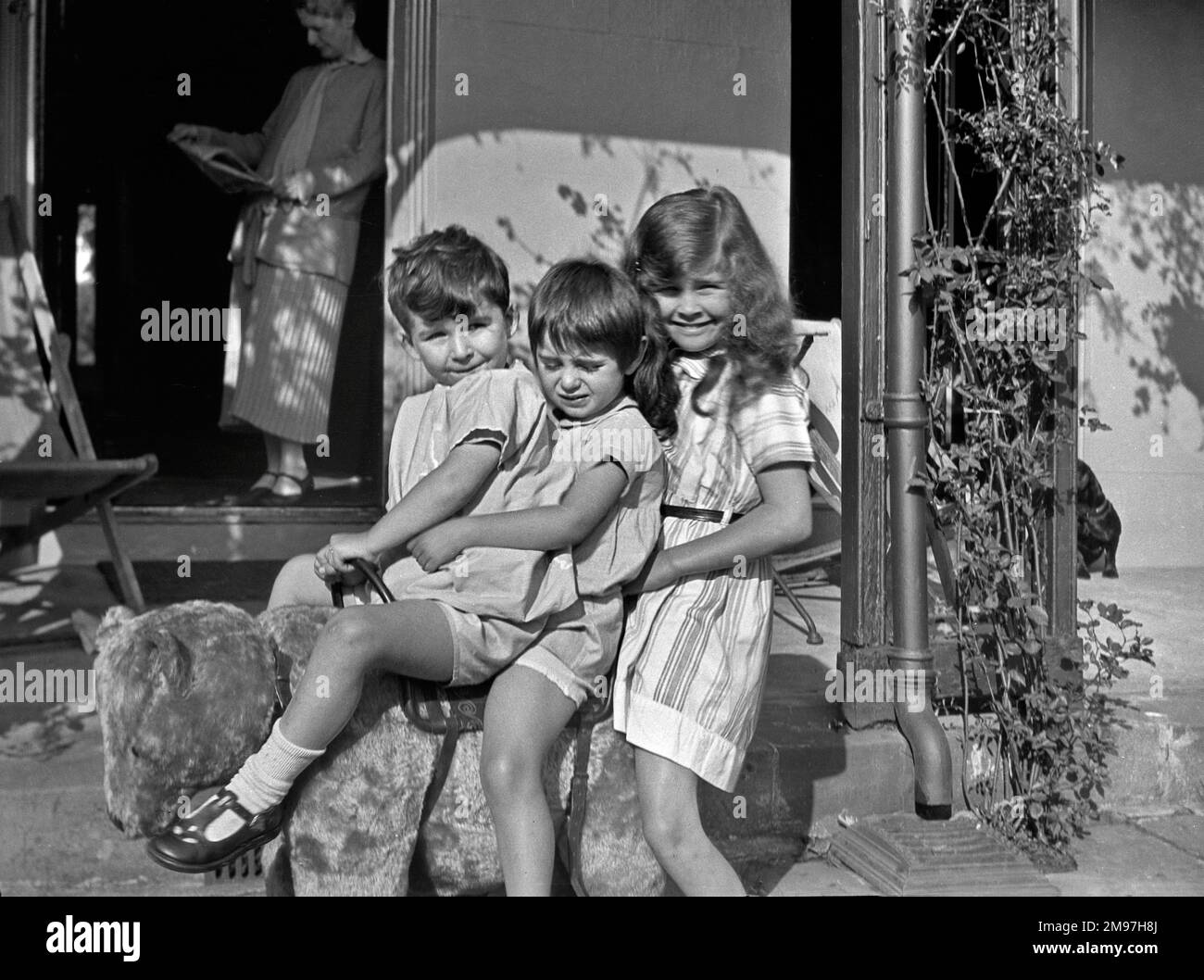 Trois enfants assis sur un ours en peluche dans un jardin, avec une femme dans une porte ouverte en arrière-plan. Banque D'Images