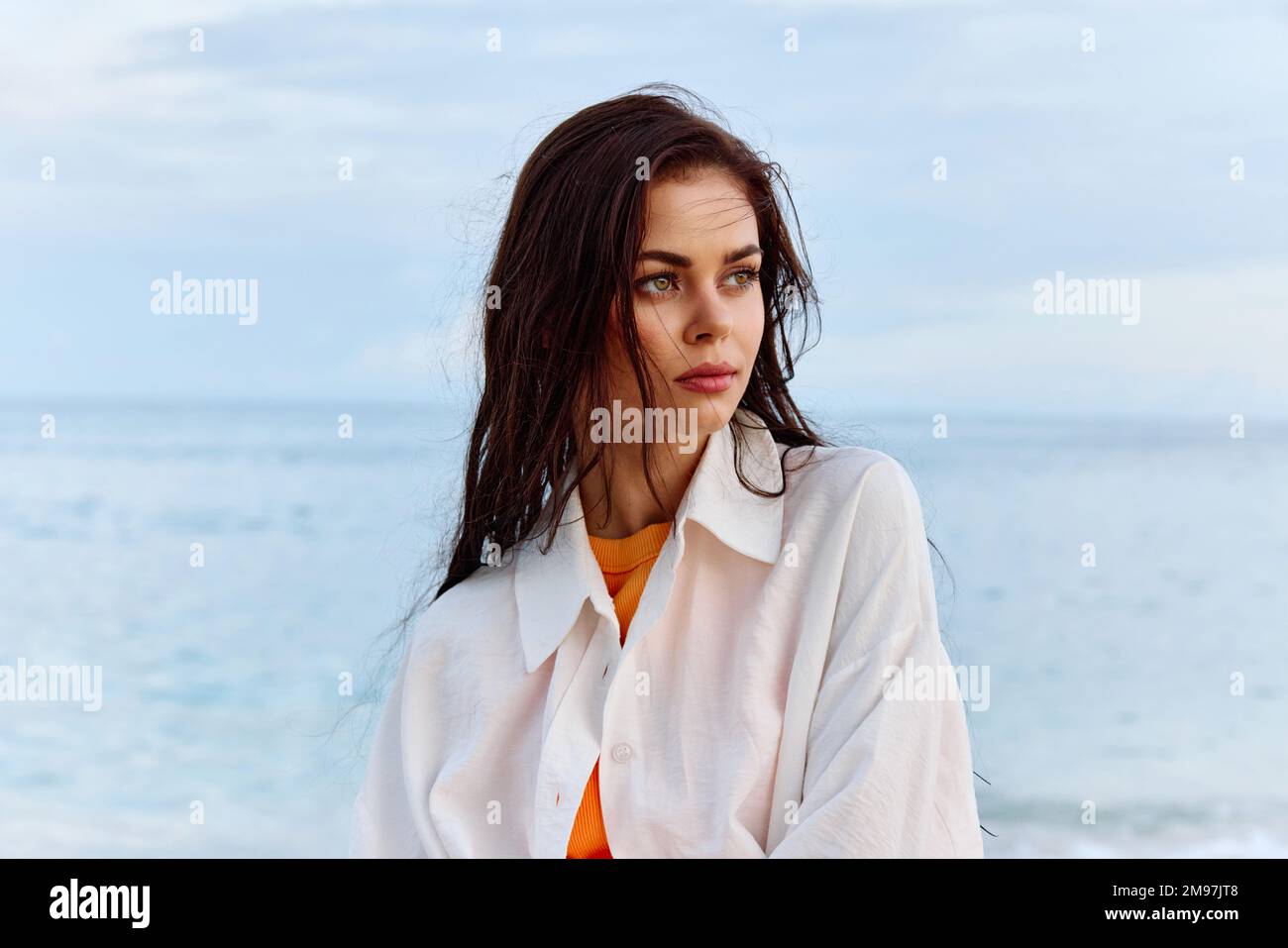 Portrait d'une belle femme pensive avec une peau bronzée dans une chemise de plage blanche avec des cheveux mouillés après avoir nagé sur la plage de l'océan coucher de soleil lumière avec des nuages Banque D'Images