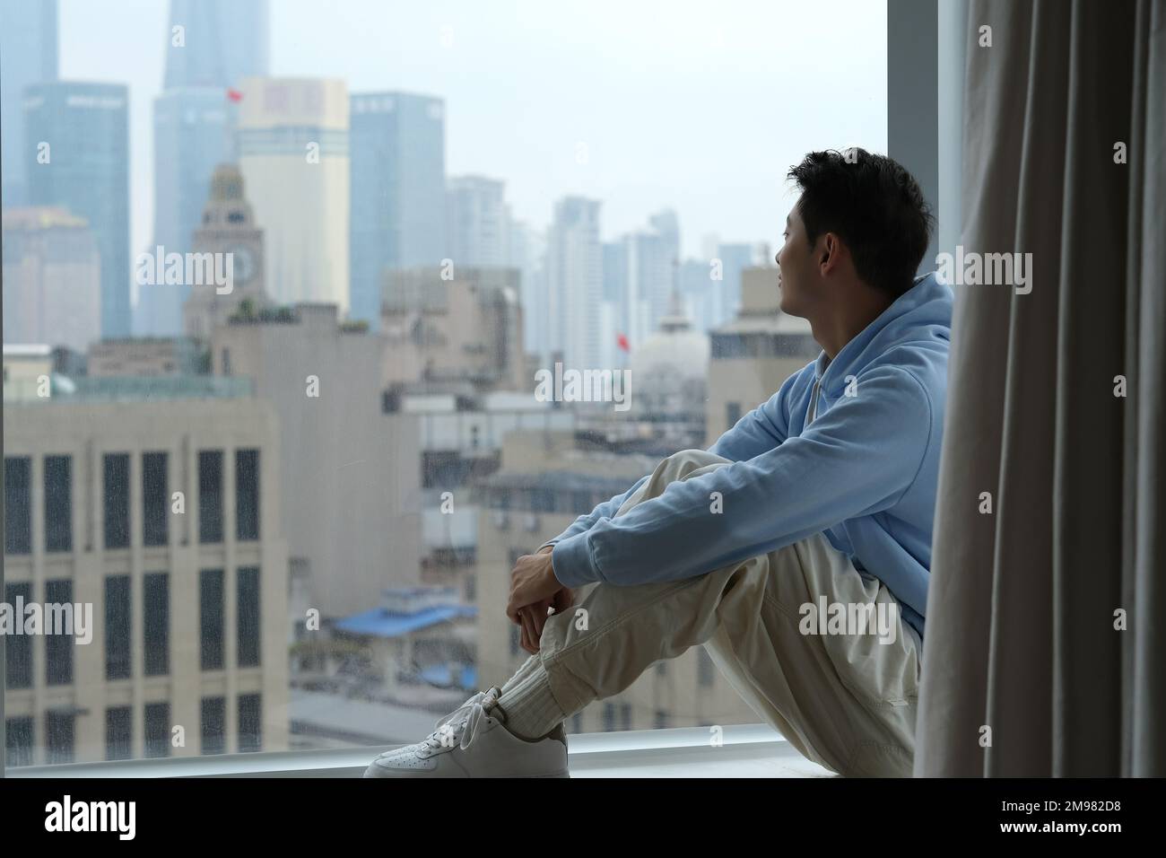 Un jeune homme chinois solitaire s'assoit au seuil de la fenêtre, regarde la fenêtre pour voir les bâtiments de la ville urbaine à Shanghai, en Chine Banque D'Images