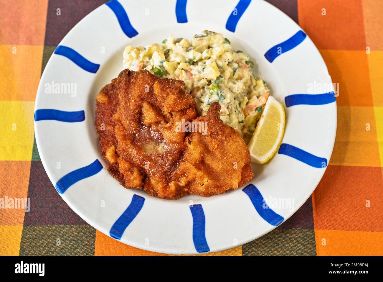 Schnitzel de porc avec salade de pommes de terre et légumes sur une assiette à rayures bleues sur une nappe orange, gros plan. Banque D'Images