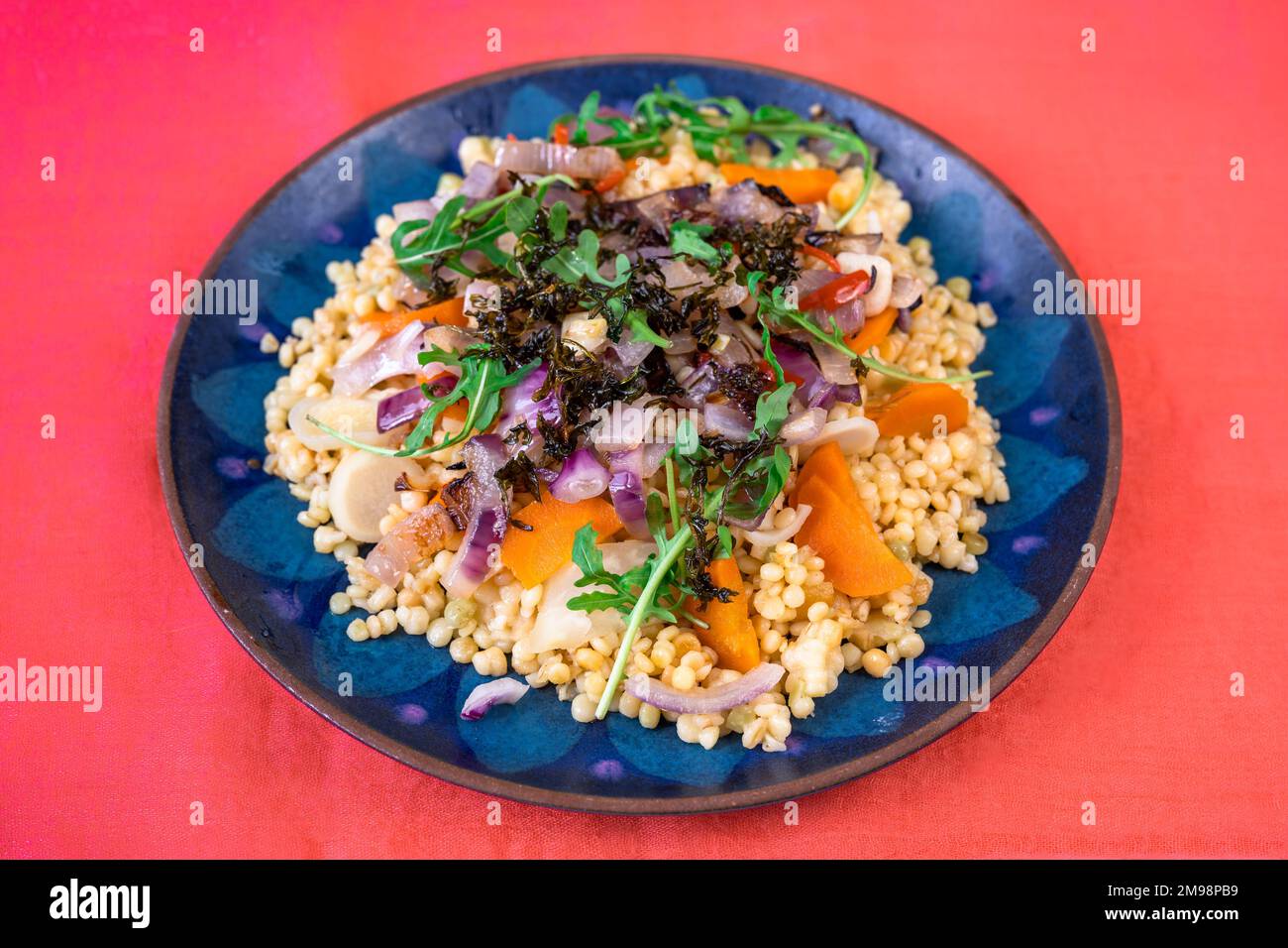 Pâtes cuites tarhona (type de pâtes européennes) avec légumes et rocola sur plaque bleue, sur fond rouge. Repas végétarien. Banque D'Images