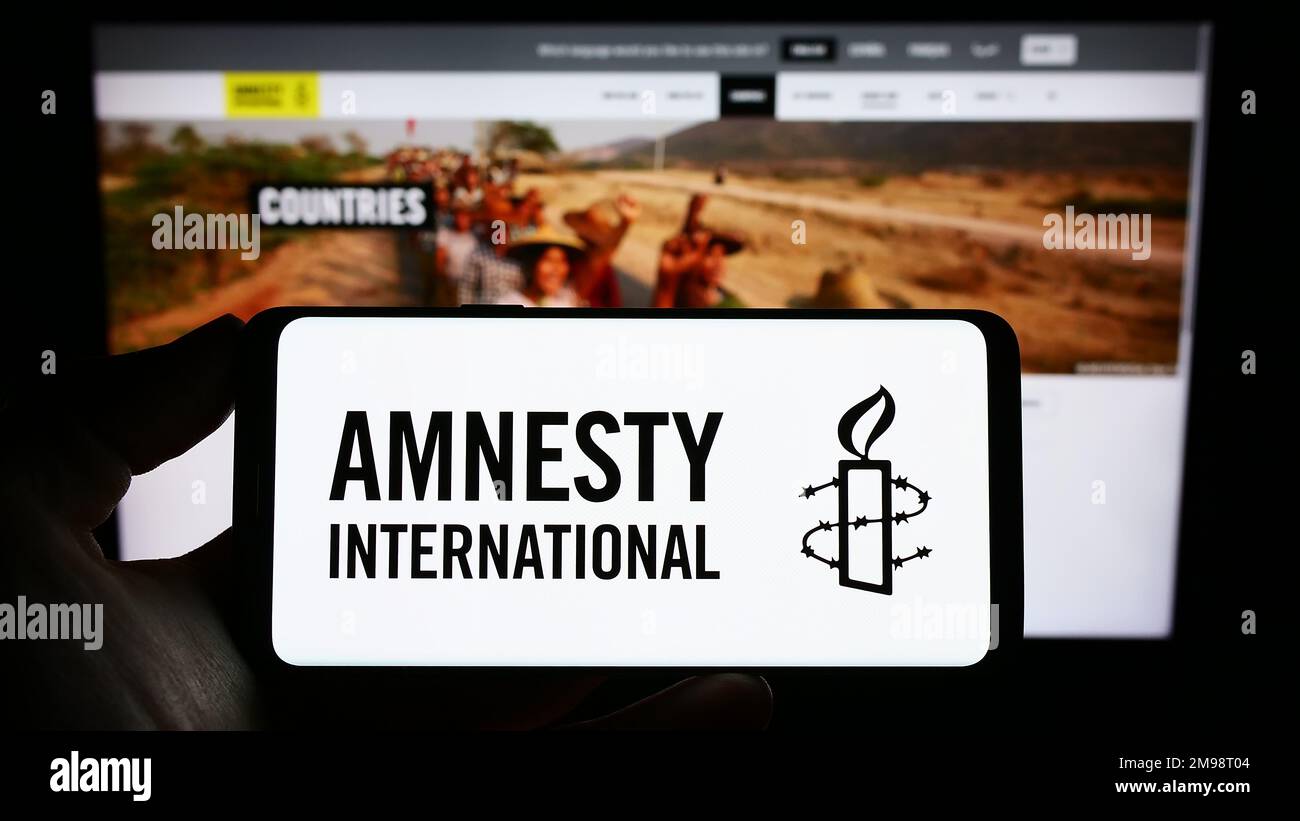 Personne tenant un téléphone portable avec le logo de l'organisation des droits de l'homme Amnesty International à l'écran en face de la page web. Mise au point sur l'affichage du téléphone. Banque D'Images