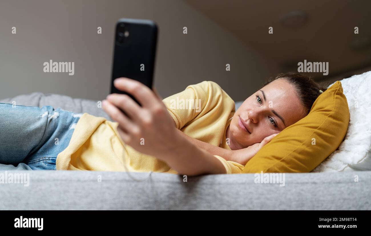 Femme avec un visage d'expression vierge allongé sur un canapé et utilisant son téléphone cellulaire. Banque D'Images