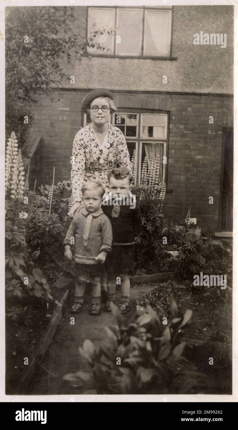 Mère (avec le fag dans sa bouche) et ses deux garçons (plutôt à face de glum), posent pour une photographie dans leur jardin soigné. Banque D'Images