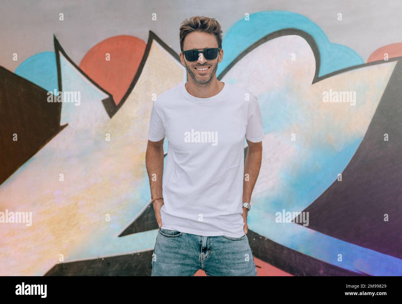 Jeune homme beau est debout sur un fond de mur de graffiti. L'homme porte un t-shirt blanc, vide de base sans logo. Banque D'Images