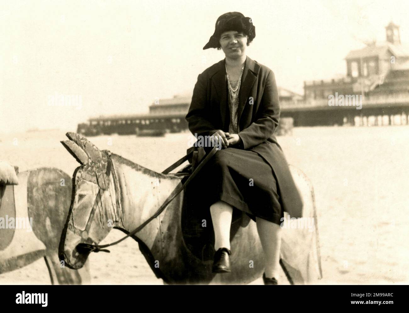 Femme perchée sur un âne en bois au bord de la mer, avec une jetée en arrière-plan (emplacement inconnu) Banque D'Images