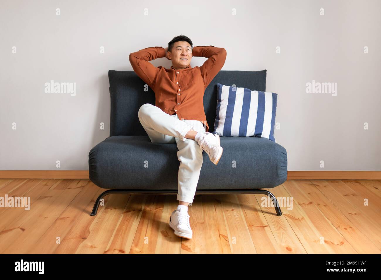 Week-end paresseux. Joyeux homme asiatique mûr se détendant avec les mains derrière la tête, assis sur un canapé au-dessus du mur blanc Banque D'Images
