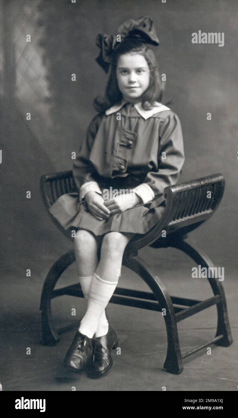 Jolie jeune fille avec un énorme noeud en velours dans ses cheveux assis pour un portrait photographique studio sur un élégant tabouret incurvé siège. Banque D'Images