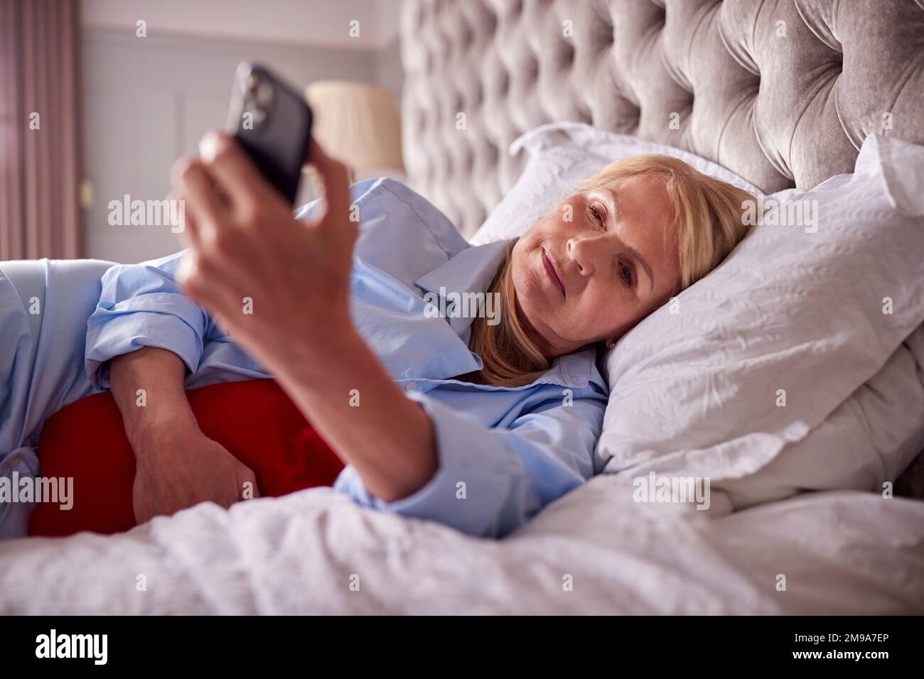 Femme mature menopausal couché au lit avec une bouteille d'eau chaude à la maison en utilisant le téléphone portable Banque D'Images